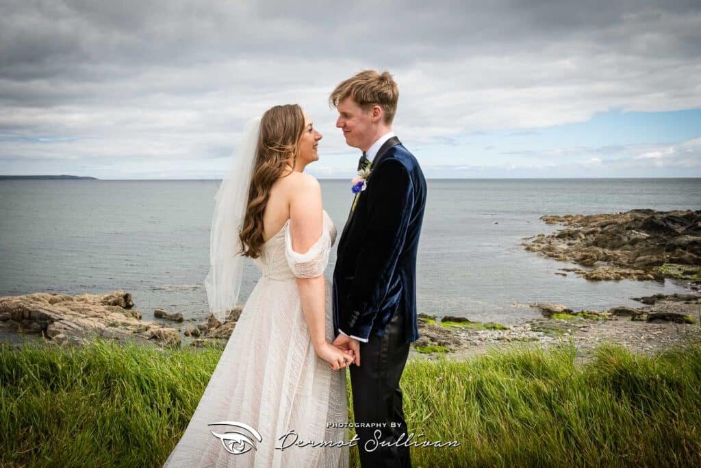 A bride and groom hold hands overlooking the sea after their outdoor wedding ceremony at Dunmore House Hotel in Cork, Ireland